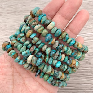 Pu&ograve; includere: Diversi braccialetti con perline di turchese. Le perline sono un mix di colori blu, verde, marrone e crema. I braccialetti sono infilati insieme e sembrano essere fatti di pietra naturale. I braccialetti sono tenuti in una mano.