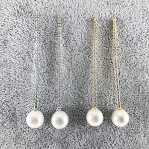 May include: Four pairs of dangle earrings with a large white pearl at the bottom. Two pairs have silver chains, and two have gold chains. The earrings are displayed on a gray, textured surface.