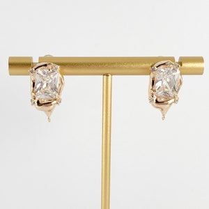 May include: A pair of gold-tone earrings with a large, clear square-shaped crystal set in a melted-looking design.