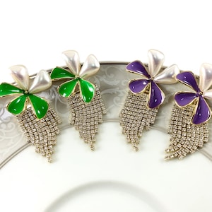 Può includere: Quattro paia di orecchini pendenti con un design a forma di fiore. I fiori sono realizzati in smalto bianco, verde e viola. Gli orecchini hanno un tono dorato e sono decorati con strass.