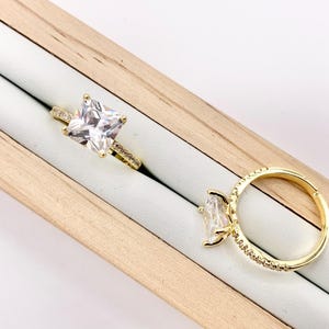 May include: Two gold rings with clear, square and oval-cut gemstones. One ring features a square-cut stone, while the other has an oval-cut stone. Both rings are set with smaller accent stones along the band. The rings are displayed in a wooden jewelry box.
