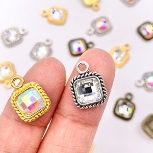 May include: A close-up of two square-shaped charms with rhinestones. One charm is silver with a clear rhinestone and the other is gold with an iridescent rhinestone. Both charms have a rope-like border.