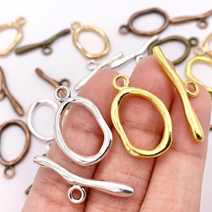 May include: A collection of metal toggle clasps in various colors, including gold, silver, and bronze. The clasps feature an oval ring and a bar with a loop. These jewelry findings are suitable for necklaces and bracelets.