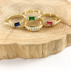 May include: Four gold rings with different colored gemstones. The rings are stacked on a wooden surface. The gemstones are green, blue, red, and clear.
