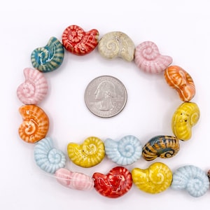 May include: A collection of multi-coloured ceramic beads shaped like spiral seashells, arranged in a circular pattern around a silver-coloured United States quarter coin for scale. The beads feature glossy finishes in shades of red, blue, yellow, orange, pink, and beige.