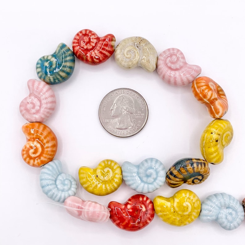 May include: A collection of multi-coloured ceramic beads shaped like spiral seashells, arranged in a circular pattern around a silver-coloured United States quarter coin for scale. The beads feature glossy finishes in shades of red, blue, yellow, orange, pink, and beige.