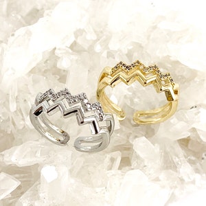 May include: Two adjustable rings with a zig-zag design and clear cubic zirconia stones. One ring is silver and the other is gold.