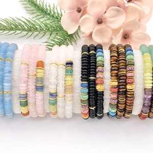 Peut inclure: Plusieurs bracelets de perles de diff&eacute;rentes couleurs, dont bleu clair, rose, blanc, noir, marron, jaune et vert. Chaque bracelet est compos&eacute; de petites perles plates s&eacute;par&eacute;es par des entretoises dor&eacute;es. Les bracelets sont pr&eacute;sent&eacute;s sur un tissu blanc.