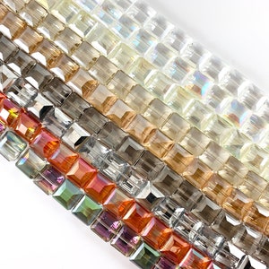 May include: Assortment of square glass beads in various colors, including clear, amber, gray, and iridescent shades. The beads are arranged in rows, showcasing their different hues and textures.