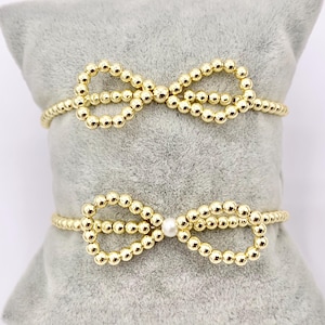 May include: Two gold beaded bracelets with a bow design. The smaller bracelet has a single white pearl accent.