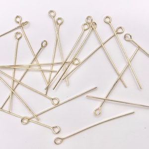 May include: A group of gold-colored head pins with a loop at the top. The pins are scattered on a white surface.