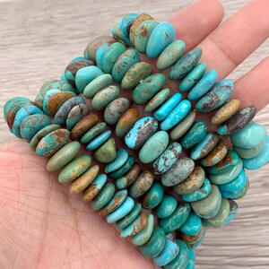 May include: A collection of turquoise gemstone beads in shades of blue, green, and brown. The oval-shaped beads are strung together, forming a textured accessory. The beads are held in a hand.