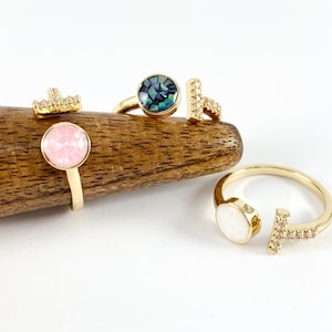 May include: A collection of gold-toned rings with various designs. One ring features a pink, faceted stone, while another has a blue-green mosaic-like stone. Other rings include a T-shaped design with small clear stones and a white stone.