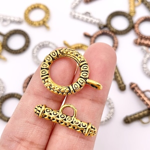 May include: Close-up of various metal toggle clasps in gold, silver, and bronze tones. The clasps feature intricate designs and are used for jewelry making. The clasps are shown in a variety of shapes and sizes.