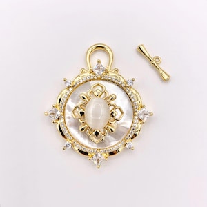 May include: A gold-toned pendant with a central oval-shaped, off-white stone, surrounded by a white mother-of-pearl background. The pendant features a decorative gold frame with clear, square and round crystals. A separate gold bar clasp is also visible.