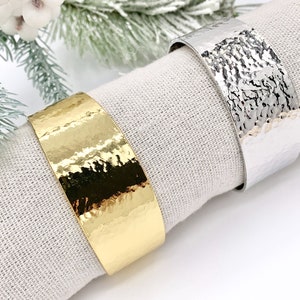 May include: Two hammered metal napkin rings, one gold and one silver, on a white linen napkin.