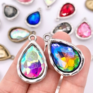 May include: Close-up of several teardrop-shaped pendants with iridescent, multicolored crystals. Each pendant has a silver-colored, textured bezel and a small loop at the top for attaching to a chain or other jewelry component. The crystals reflect a rainbow of colors.