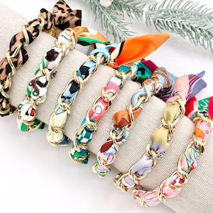 May include: A collection of fabric-covered hair accessories with gold chain accents. The scrunchies feature various patterns, including animal print, floral, and geometric designs. The scrunchies are arranged on a neutral-colored surface.
