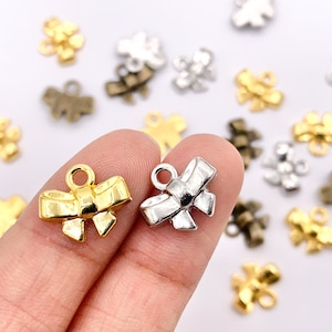 May include: Two gold and silver bow charms with a loop for attaching to jewelry or crafts. The charms are on a finger, with other charms scattered around.