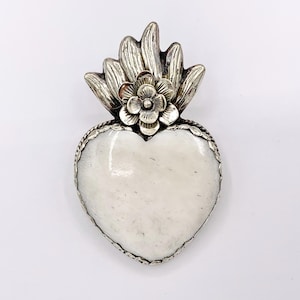 May include: A silver-toned pendant in the shape of a heart, featuring a white stone center. The pendant is adorned with a floral design at the top and a decorative border. The heart-shaped pendant is a unique piece of jewelry.