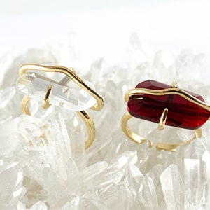 May include: Two gold rings with clear and red gemstone settings. The rings are set on a bed of white crystals.