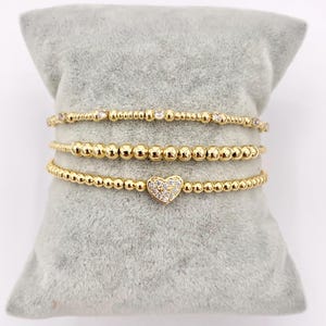 May include: Three gold beaded bracelets. One bracelet features small clear stones, another has solid gold beads, and the third has a heart-shaped charm encrusted with clear stones. The bracelets are displayed on a gray pillow.