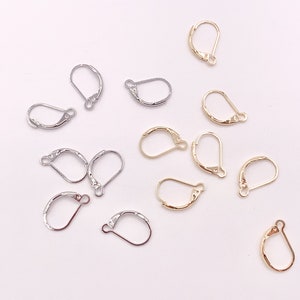May include: An assortment of leverback earring findings in gold and silver tones. The findings are arranged on a white surface, showcasing their curved design and small loop for attaching earrings.