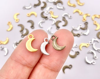 May include: A close-up of three crescent moon charms. The charms are made of metal and have a gold, silver, and antique bronze finish. The charms are being held by a hand.