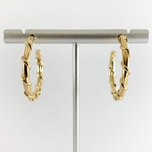 May include: Gold hoop earrings with a twisted design. The earrings are displayed on a silver stand.