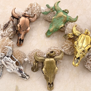 May include: Five different metal bull skull charms in various finishes, including silver, gold, and green. The charms are displayed on a white background with a soft, textured surface.