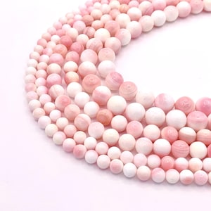 May include: A collection of smooth, spherical beads arranged in a curved formation. The beads feature a gradient of soft pink and white hues, with some displaying subtle, natural marbled patterns and a polished, glossy finish throughout the set.