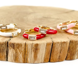 May include: Four pairs of gold hoop earrings with colorful enamel and clear cubic zirconia stones. The earrings are arranged on a wooden surface.