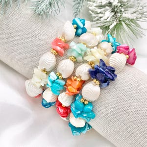 May include: A colorful bracelet featuring a variety of seashells and small, colorful, polished stones. The bracelet includes white, blue, pink, orange, and green elements, with gold-colored beads between each section. The bracelet is resting on a light gray surface.