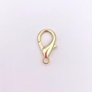 May include: A gold-toned metal lobster claw clasp featuring a smooth, polished finish with a spring-loaded mechanism and a small circular loop at the base for attachment, designed for jewelry making and securing various accessories.