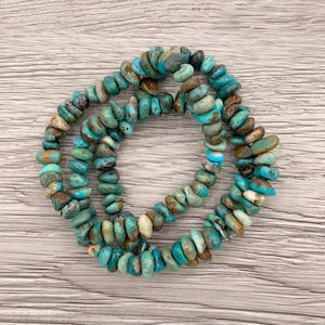 Pu&ograve; includere: Tre braccialetti con perline di schegge di turchese. Le perline sono un mix di colori blu, verde, marrone e crema. I braccialetti sono disposti in un motivo circolare su una superficie di legno chiaro.