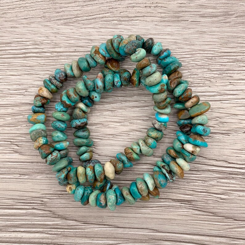 Pu&ograve; includere: Tre braccialetti con perline di schegge di turchese. Le perline sono un mix di colori blu, verde, marrone e crema. I braccialetti sono disposti in un motivo circolare su una superficie di legno chiaro.