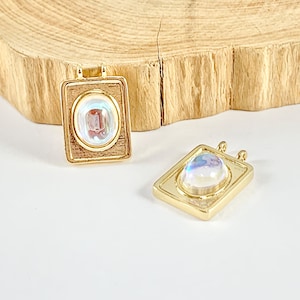 May include: Two gold-toned pendants with oval moonstone gems set in a square frame. The pendants have a textured surface and are ready to be added to a necklace or bracelet.