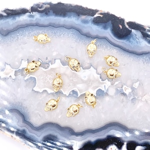 May include: A collection of gold-toned skull charms on a white and gray agate background. The charms are small and detailed, with a three-dimensional design.