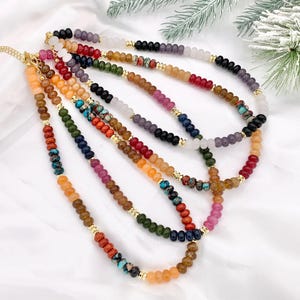 May include: A collection of colorful beaded necklaces. Each necklace features a variety of gemstone beads in shades of red, orange, yellow, green, blue, purple, and white. Gold-colored metal accents separate the bead sections. The necklaces are displayed on a white surface.
