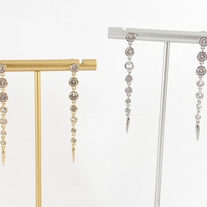 May include: Two pairs of dangle earrings with a row of clear round stones and a pointed tip. The earrings on the left are gold and the earrings on the right are silver.