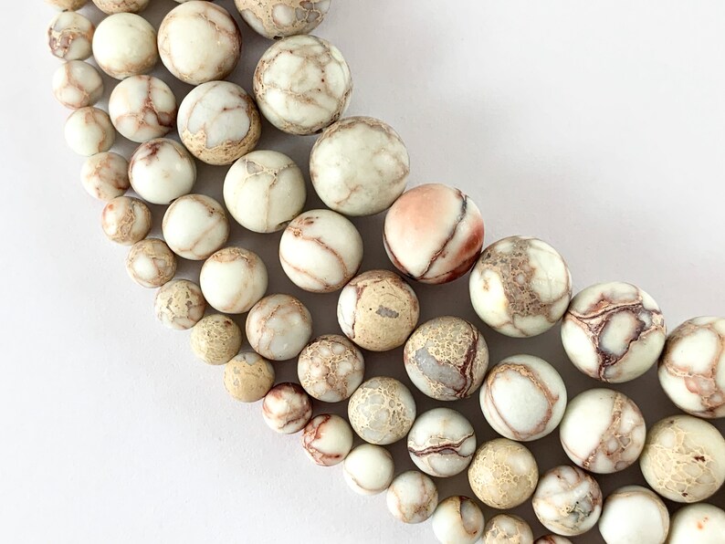 May include: A strand of white, brown, and beige jasper beads with a marbled pattern. The beads are round and smooth, and they are arranged in a graduated size from smallest to largest.