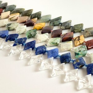 May include: A strand of gemstone beads in various colors and patterns, including green, blue, red, and brown. The beads are faceted and have a square shape. The beads are strung on a clear thread.
