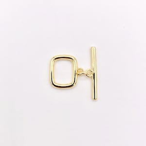 May include: A gold-colored toggle clasp, featuring a rectangular loop and a straight bar. The clasp is connected by small rings, designed for jewelry making or crafting projects. The hardware is set against a white background.