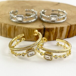 May include: A pair of gold hoop earrings with a double row of clear cubic zirconia stones. The earrings have a rectangular stone in the center of each hoop.