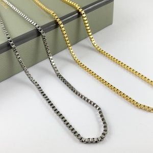 May include: Three delicate necklaces with square links. One is gunmetal, and the other two are gold. The necklaces are displayed on a white surface with a gray box in the background. Jewelry.