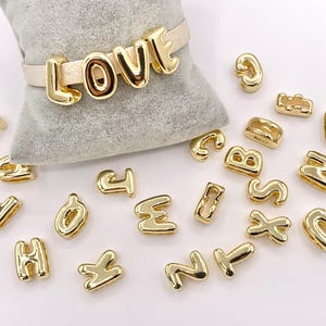 May include: Gold-colored metal letter beads and a bracelet spelling "LOVE" on a gray pillow. Individual letters are scattered around the pillow. The bracelet has a light tan band. The letters are in a bold, rounded font.