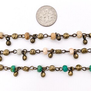 May include: Three strands of chain with beads and charms. The top strand has brown and beige beads, the middle strand has white and gray beads, and the bottom strand has green beads. All strands have small, round, bronze charms.