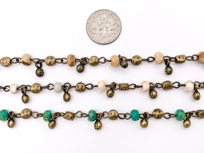 May include: Three strands of chain with beads and charms. The top strand has brown and beige beads, the middle strand has white and gray beads, and the bottom strand has green beads. All strands have small, round, bronze charms.