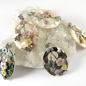 May include: Four oval-shaped pendants made of iridescent mother-of-pearl with floral designs. The pendants feature delicate silver branches with pink and yellow flowers.