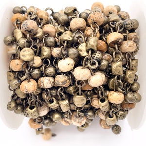 May include: A close-up of a chain made of brown and beige stone beads and antique gold metal links. The chain is wound around a white spool.